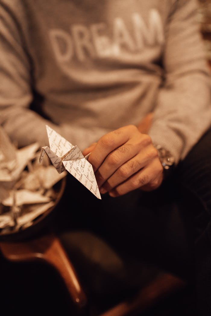 Unrecognizable male in casual clothes sitting with traditional decorative paper crane in hand near bowl full of orizurus on blurred background